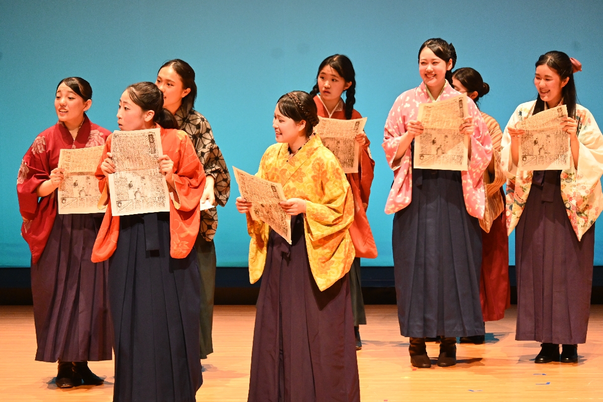 物語終盤、笑顔を浮かべる女学生たち。新聞を手にしている