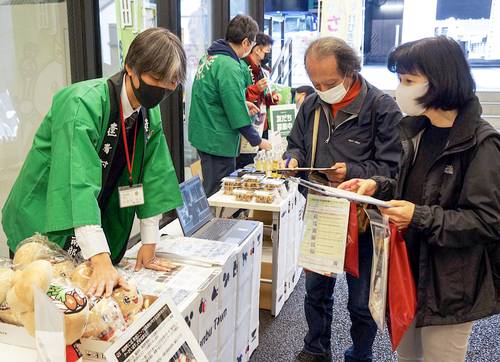 自治体のブースで特産などの説明を聞きながらクイズに答える来場者(右側)=5日、東京・内幸町