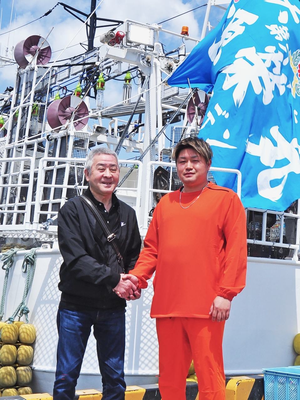 「第67源栄丸」の前でバトンタッチの握手を交わす大橋社さん(左)と米倉龍也さん=5日、八戸港